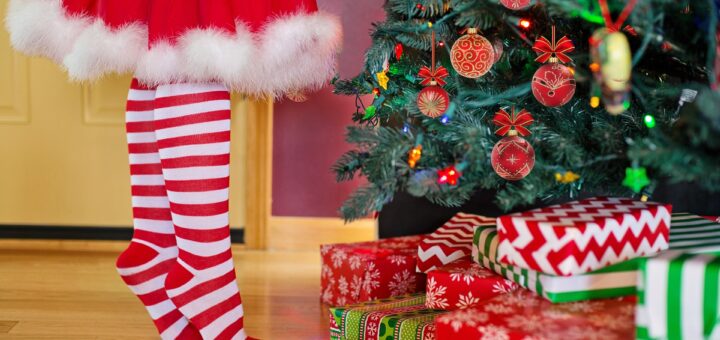 Üppig geschmückter Weihnachtsbaum und ein Mädchen im Weihnachtskostüm, das von der Taille abwärts in rot-weiß-gestreiften Kniestrümpfen zu sehen ist